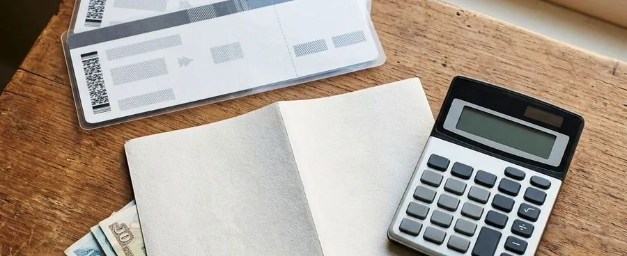 A travel itinerary, calculator, and card laid out beside a stack of cash