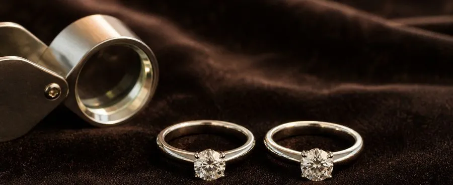 Two nearly identical diamond solitaire rings on a velvet pad beside a jeweler's loupe.