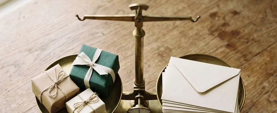 A small balance scale weighing wrapped gift boxes against a stack of plain envelopes