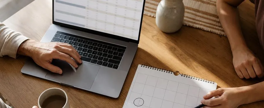 A couple's budget spreadsheet and wall calendar on a kitchen table, timing a honeymoon departure date
